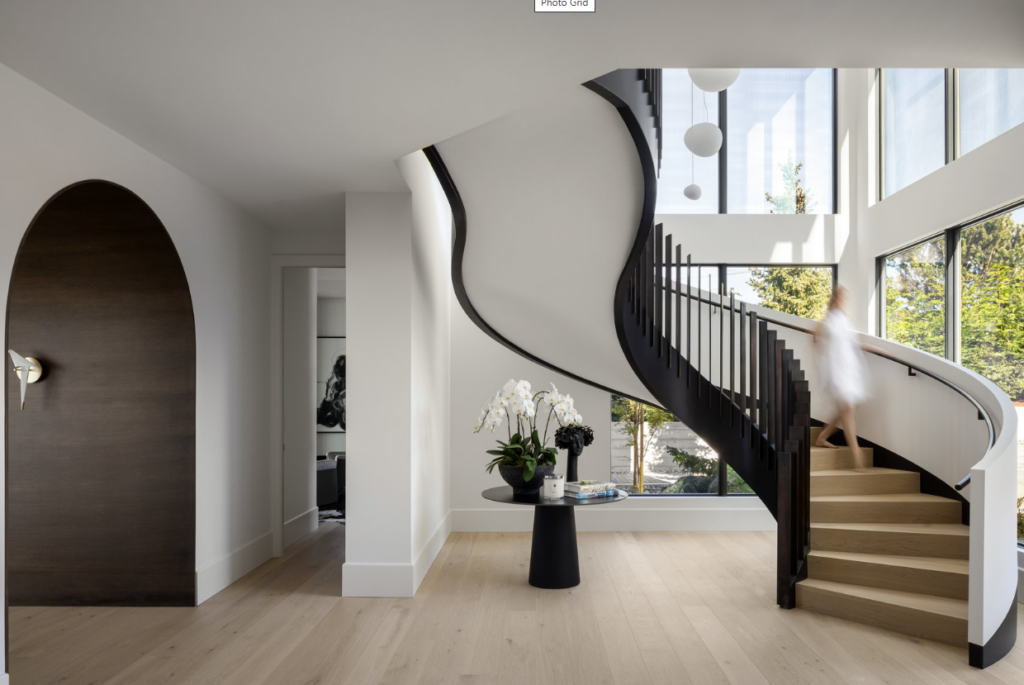 Modern interior entryway at 4040 White Rock Street in Saanich, British Columbia, showcasing a dramatic spiral staircase with a curved black handrail and wooden steps, light hardwood flooring, a dark arched accent door, and bright floor-to-ceiling windows.