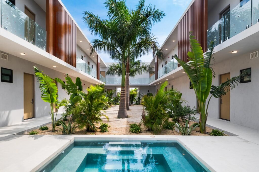 Central courtyard and swimming pool at The Retreat in Grace Bay, Turks and Caicos, showcasing modern two-story condo-hotel buildings with glass-paneled balconies, slatted wood panel accents, lush palm trees, and tropical landscaping.