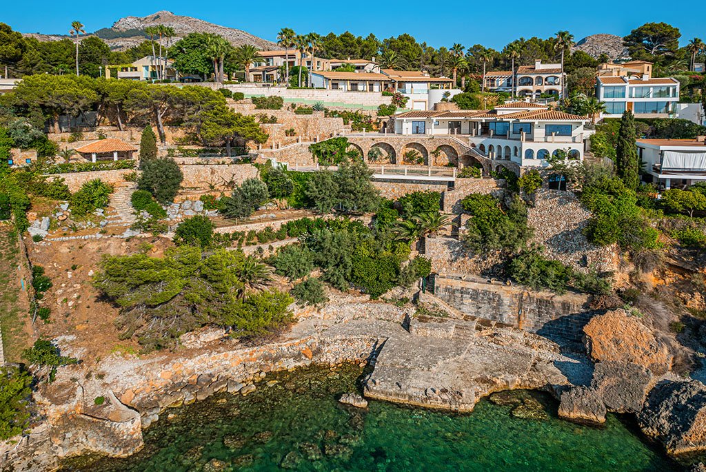 An aerial view of a terraced cliffside luxury villa in Bonaire, Mallorca, featuring cascading stone-walled gardens that lead down to a private jetty with direct access to crystal-clear turquoise sea water. The multi-level white residence sits at the top of the hillside plot surrounded by Mediterranean pine trees and rocky coastline under a clear blue sky.