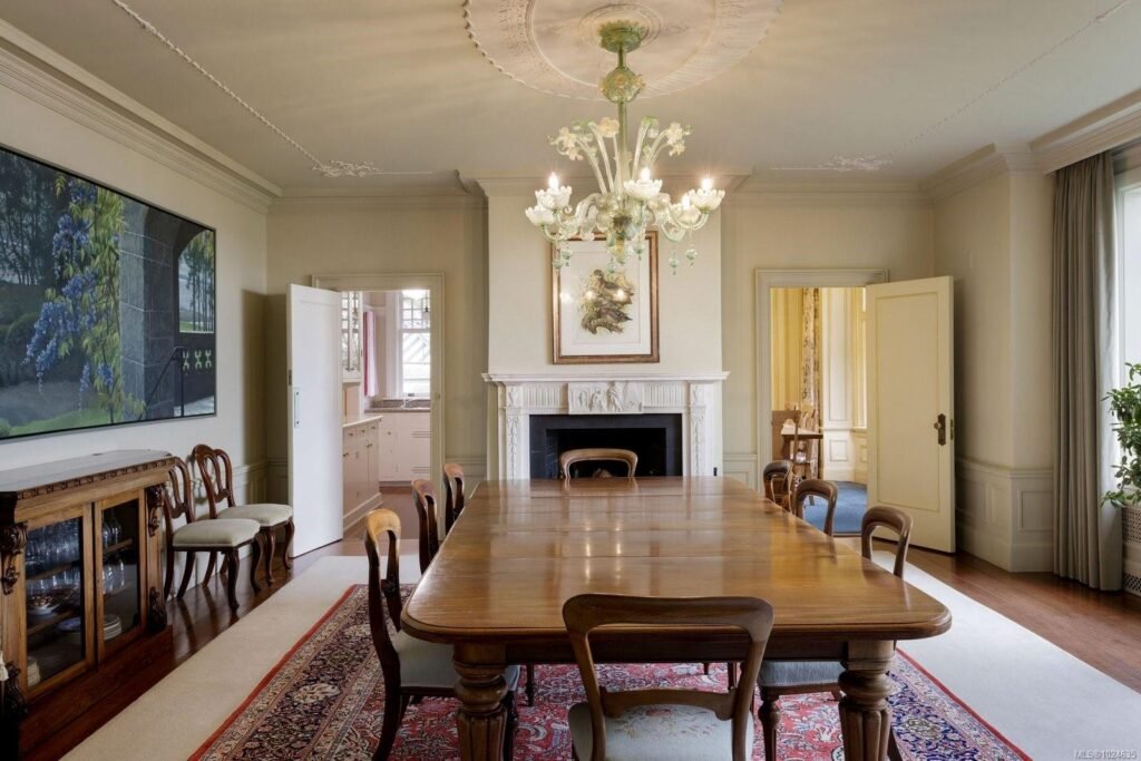 Formal dining room interior of the historic Riffington estate at 3175 Beach Drive, Oak Bay, British Columbia, featuring a large antique wooden dining table, a classic white fireplace, an ornate glass chandelier, and elegant crown molding.