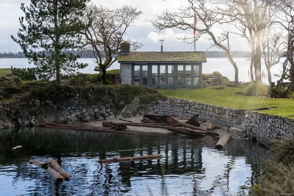 Seaside waterfront pavilion and stone-walled private cove at the historic Riffington estate at 3175 Beach Drive, Oak Bay, British Columbia, showing calm ocean waters, natural logs along the shoreline, and mature trees.