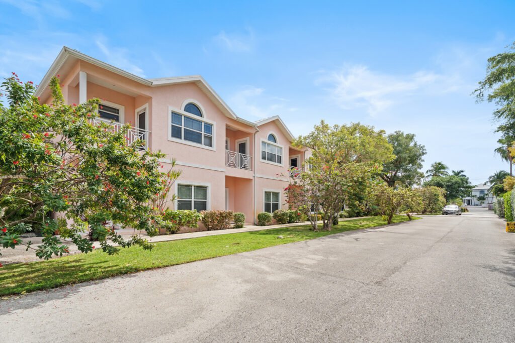 Exterior view of a pink townhouse complex at Southern Skies in South Sound, Grand Cayman, featuring multi-story residential units, manicured green lawn, lush tropical trees, and a paved neighborhood driveway under a blue sky.