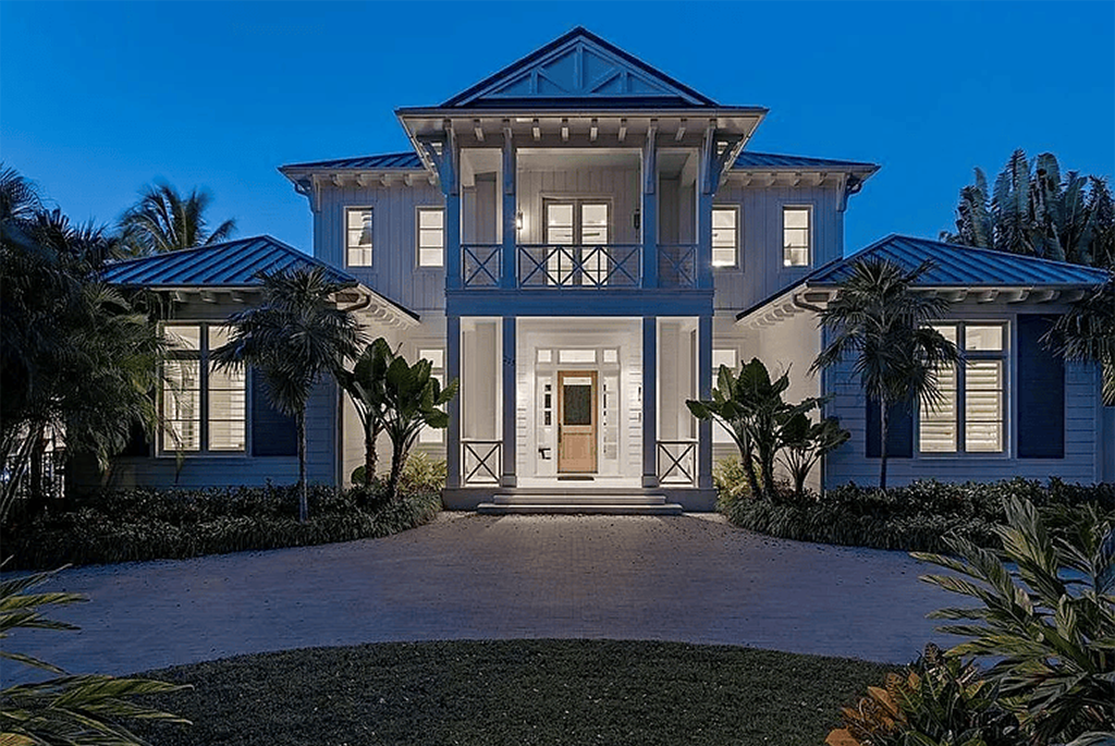 Twilight exterior view of the luxury coastal estate at 225 4th Avenue N in Olde Naples, Florida, showcasing a symmetrical two-story main residence with illuminated windows, a grand columned entryway, a second-floor balcony, a brick-paved driveway, and lush tropical landscaping under a deep blue evening sky.