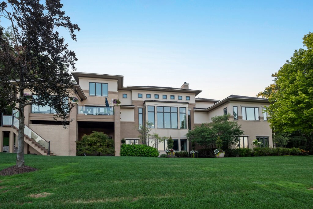 Rear exterior view of the luxury lakefront estate at 4969 Elmgate Drive, Orchard Lake, Michigan, showcasing a large brick contemporary mansion with sprawling green lawns and expansive windows overlooking Upper Straits Lake.