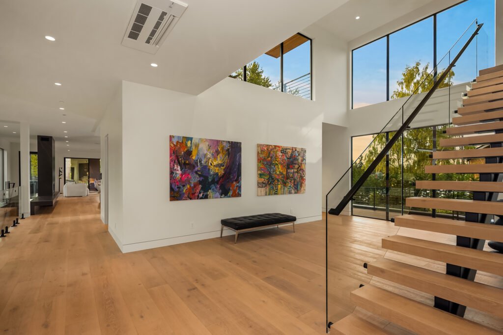 Spacious entry foyer and floating staircase at The 8528 in Clyde Hill, Seattle, featuring light hardwood floors, a modern black leather bench, large glass windows, and vibrant abstract artwork curation.