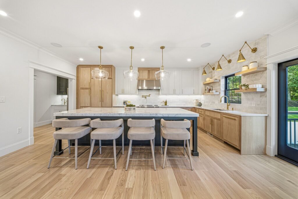 The designer chef's kitchen inside 211 High Rock Street, a custom-built luxury home in Massachusetts featuring a large marble island with seating, natural wood cabinetry, and elegant glass pendant lighting.