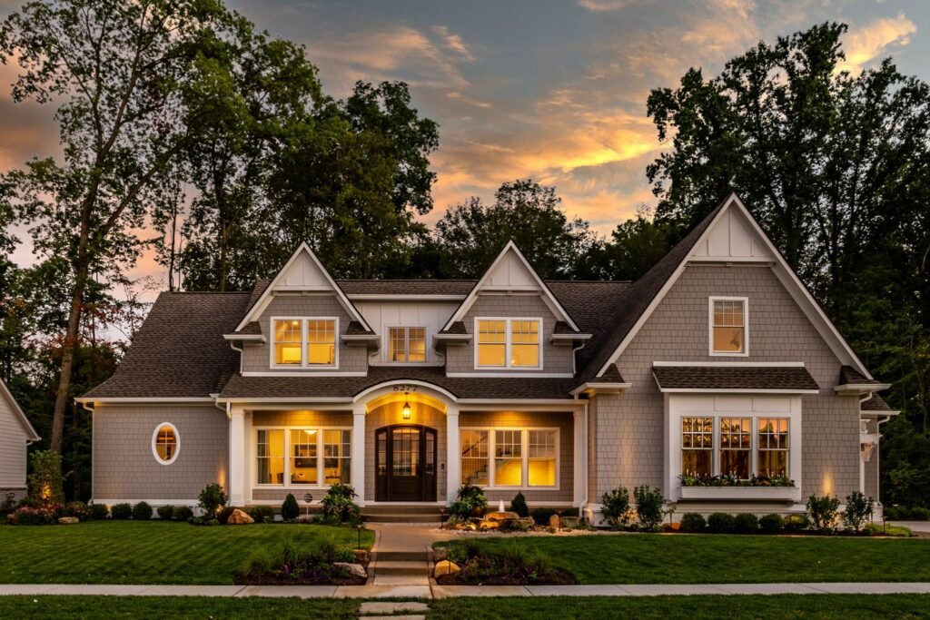 The exterior front facade of 8277 Keeneland Road, a New England shingle-style custom home by Gradison Design Build, featuring warm interior lighting and a manicured lawn at sunset in Indiana.