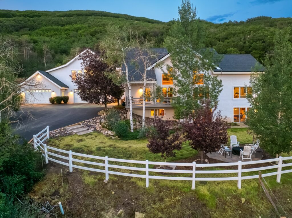 The exterior front view of 26000 High Ridge Drive, a luxury mountain estate in Steamboat Springs, Colorado, showcasing the expansive home, private driveway, white fencing, and surrounding green hills at twilight.