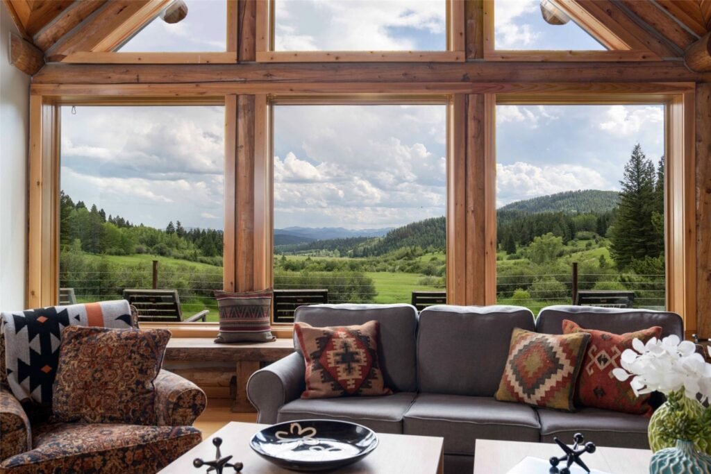 The vaulted great room inside 14200 Flaming Arrow Road, an Alpine Log Homes mountain retreat in Bozeman, Montana, showcasing a large floor-to-ceiling window wall framing sweeping views of the Bridger Canyon meadows and hills.