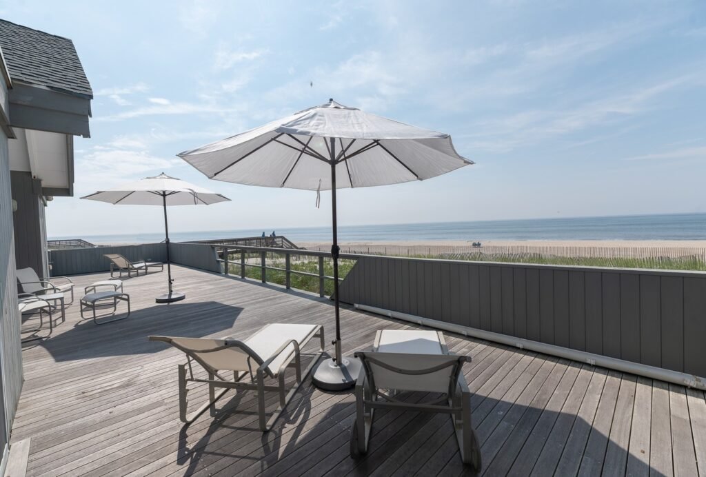 The expansive oceanfront front deck at 59 Gale Avenue, a luxury beach home in the Seaview community of Fire Island, New York, featuring lounge chairs, patio umbrellas, and sweeping views of the sandy beach and ocean.