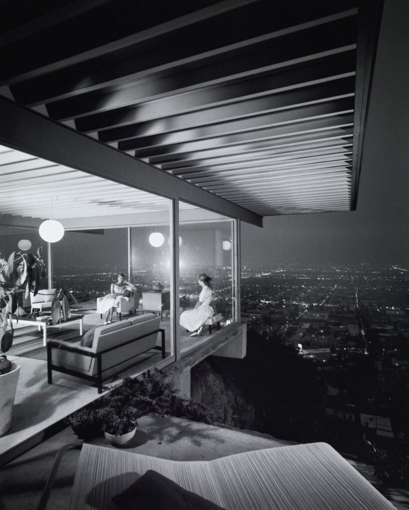 The legendary 1960 black and white photograph of the Stahl House Case Study House 22 by Julius Shulman showing two women sitting inside a glass-walled living area overlooking the night skyline of Los Angeles.