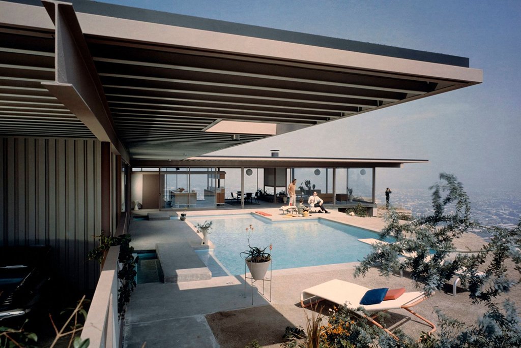 Vintage color photograph of the Stahl House Case Study House 22 outdoor swimming pool deck showing the flat steel roof beam structure and people relaxing on patio furniture.