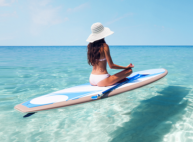 A woman in a white bikini and sun hat sitting cross-legged in a meditative pose on a paddleboard on the clear, calm turquoise water of Seven Mile Beach.