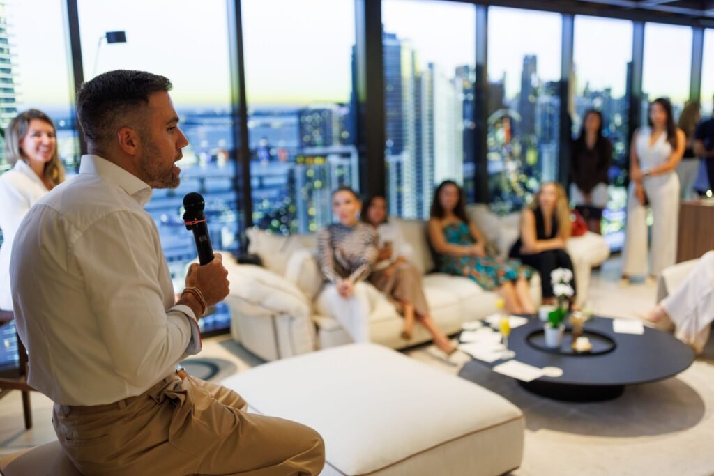 Kane Sarhan, Co-Founder of The Well, speaking into a microphone during the luxury residential launch event for HQ Residences Miami, addressing a seated audience inside a high-rise showroom with floor-to-ceiling windows overlooking the Miami skyline at twilight.