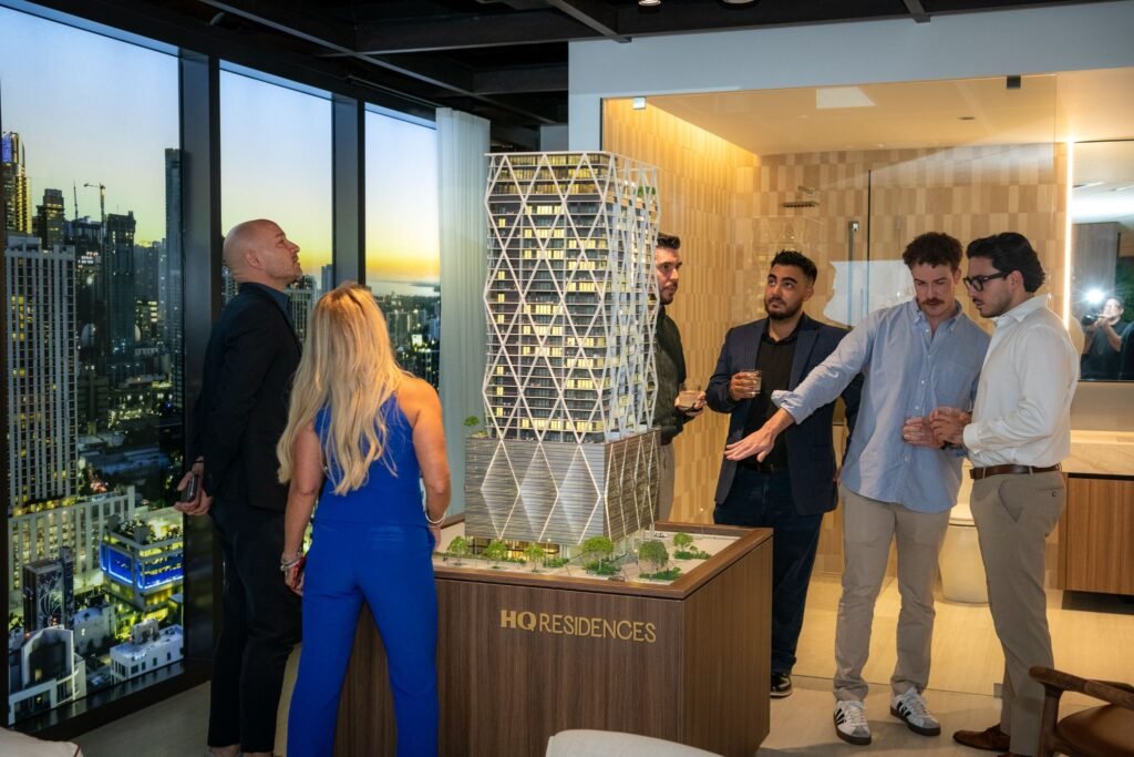 Guests gathering around the illuminated HQ Residences Miami architectural scale model during the evening social grand opening event inside a luxury sales showroom overlooking the Miami city skyline at twilight.