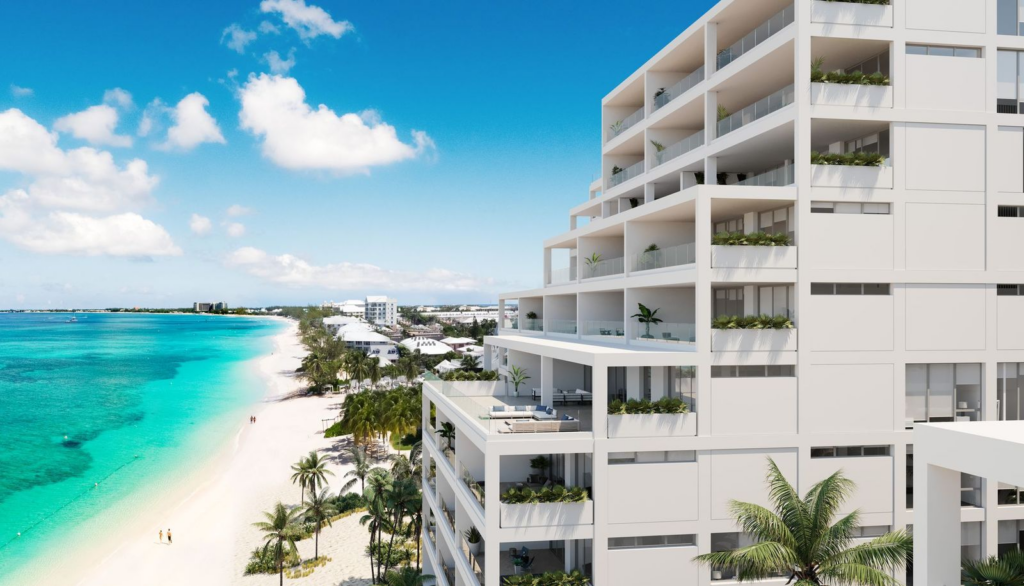 A modern, multi-story white luxury residential building featuring terraced balconies with glass railings overlooking the turquoise water and white sands of Seven Mile Beach.