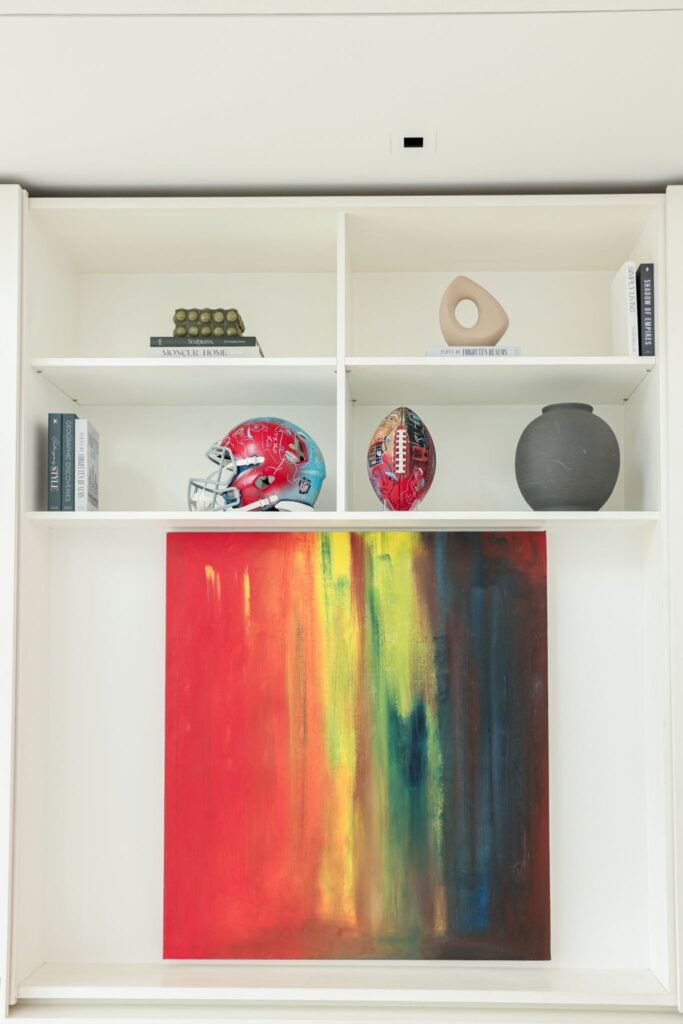 Customized luxury staging display featuring a vibrant red and yellow abstract painting below built-in white shelves styled with a painted football helmet, custom-art football, decorative vases, and books.