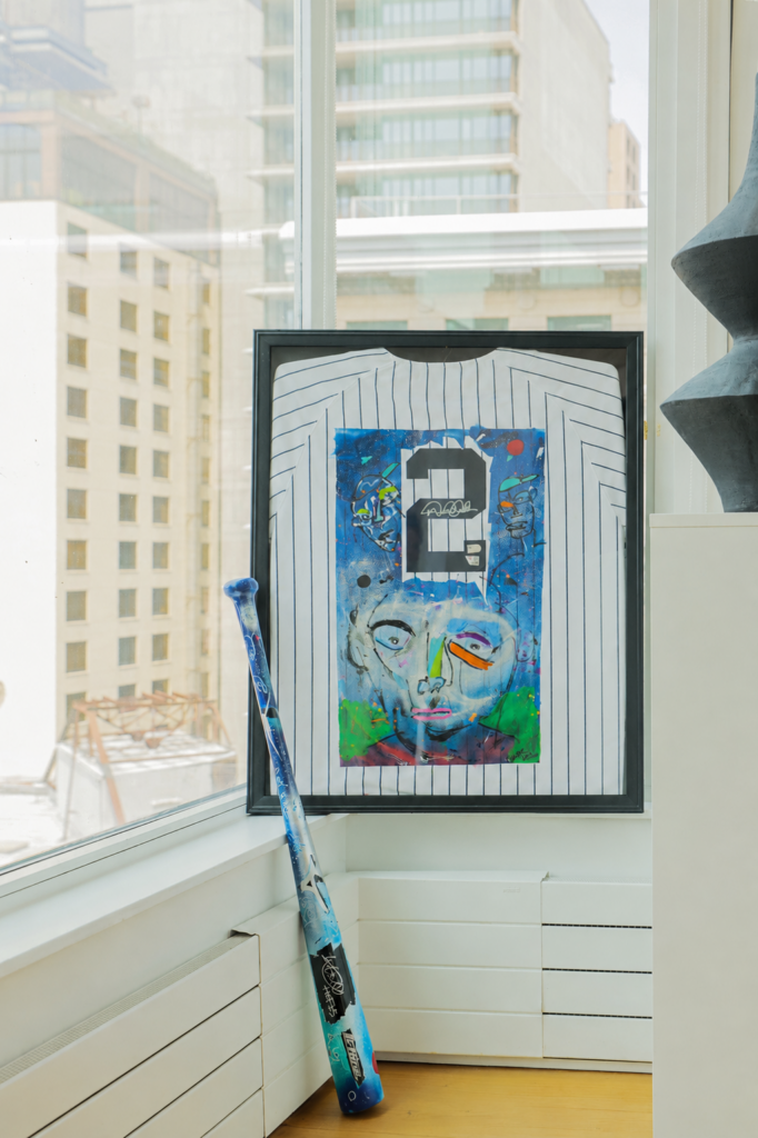A framed New York Yankees number 2 jersey featuring customized contemporary street art alongside a painted baseball bat in a luxury high-rise window alcove.