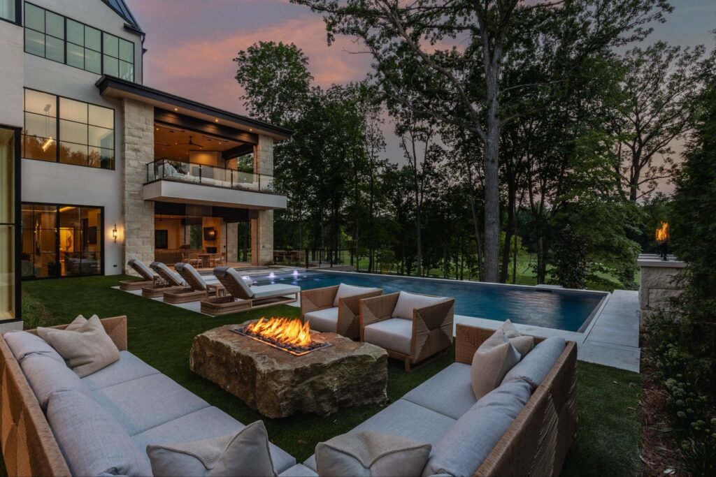 Luxury backyard patio and infinity pool at "The Reverie" estate in Zionsville, Indiana, featuring an outdoor fireplace and lounge seating at dusk