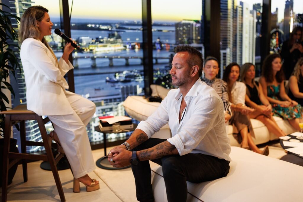 A guided group meditation session during the daytime launch event for HQ Residences Miami, featuring attendees sitting with eyes closed while a speaker in a white suit addresses the room from a stool against a panoramic window overlooking the Miami skyline and Biscayne Bay.