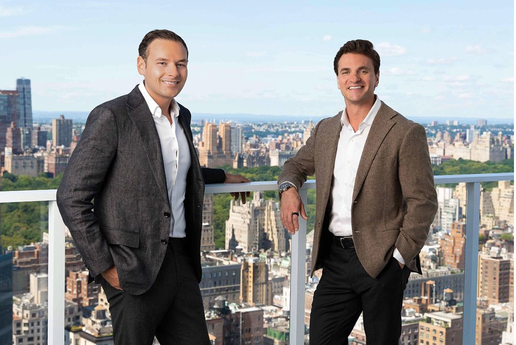 Real estate agents Michael Biryla and John Antretter of The Agency New York standing on a high-rise balcony at The Elysia overlooking the Manhattan skyline.